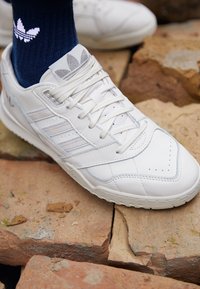 Baskets en cuir blanc avec des accents texturés, lacets plats, perforations et un contour bleu clair. Une chaussette bleue avec un logo est également visible.