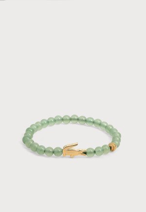 Bracelet vert à perles avec un pendentif en forme de crocodile doré et une petite perle dorée sur fond blanc.