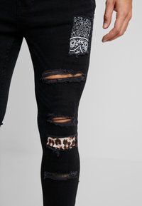 Jeans negros desgastados con secciones rasgadas, un parche con estampado de bandana y un acento de estampado de leopardo, que destacan diversas texturas y patrones.