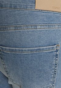 Jeans de ganga azul claro com um bolso na parte de trás e costura visível. Um retalho de couro bege com o logótipo "ESPRIT" está localizado na cintura.