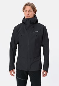 Giacca impermeabile nera con colletto alto, tasche frontali con zip, cappuccio regolabile e logo sul lato sinistro del petto. Texture liscia e opaca.