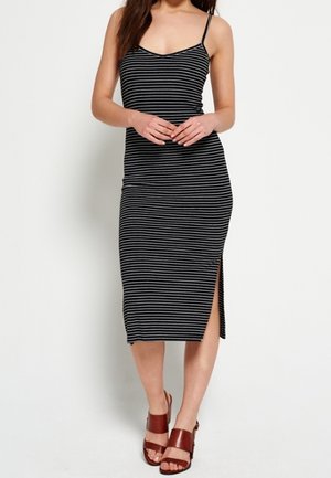 Kobieta w czarno-białej, bezrękawnikowej sukience midi w paski z bocznym rozcięciem i brązowych sandałach na obcasie z odkrytymi palcami, stojąca na białym tle.