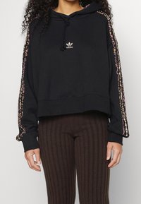 Felpa nera corta con coulisse, caratterizzata da strisce multicolori a stampa leopardata lungo le maniche e un logo dorato Adidas sul petto.