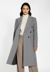 Grå dobbelbrystet frakke med struktureret overflade, klapkrave, sidelommer og sorte knapper, parret med en elfenbensfarvet turtleneck og beige bukser.