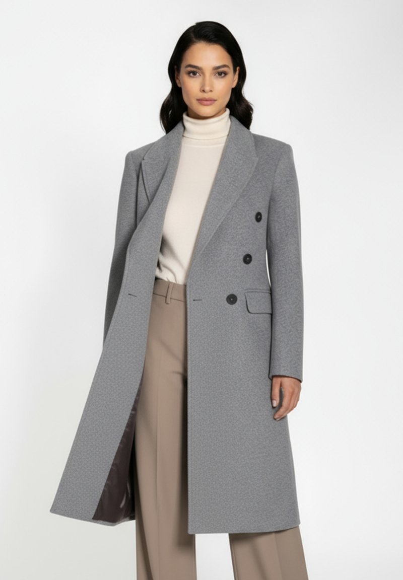 Grå dobbelbrystet frakke med struktureret overflade, klapkrave, sidelommer og sorte knapper, parret med en elfenbensfarvet turtleneck og beige bukser.