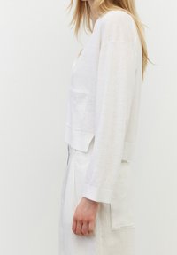 Biały krótki top z długim rękawem, głębokim dekoltem, bocznym rozcięciem i przednią kieszenią, wykonany z lekkiego lnu. Miękka tekstura.