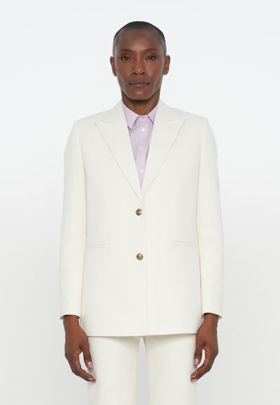 Blazer bianco su misura con due bottoni, revers a punta e tasche laterali. Indossato sopra una camicia a righe viola chiaro. Tessuto testurizzato.