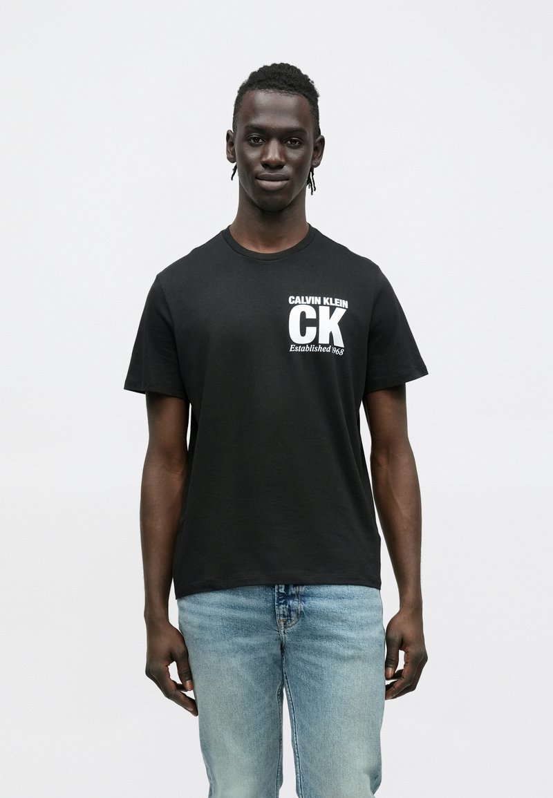 Czarny bawełniany T-shirt z białymi logo "CALVIN KLEIN" i "CK" na lewej części klatki piersiowej, zestawiony z jasnoniebieskimi dżinsami.