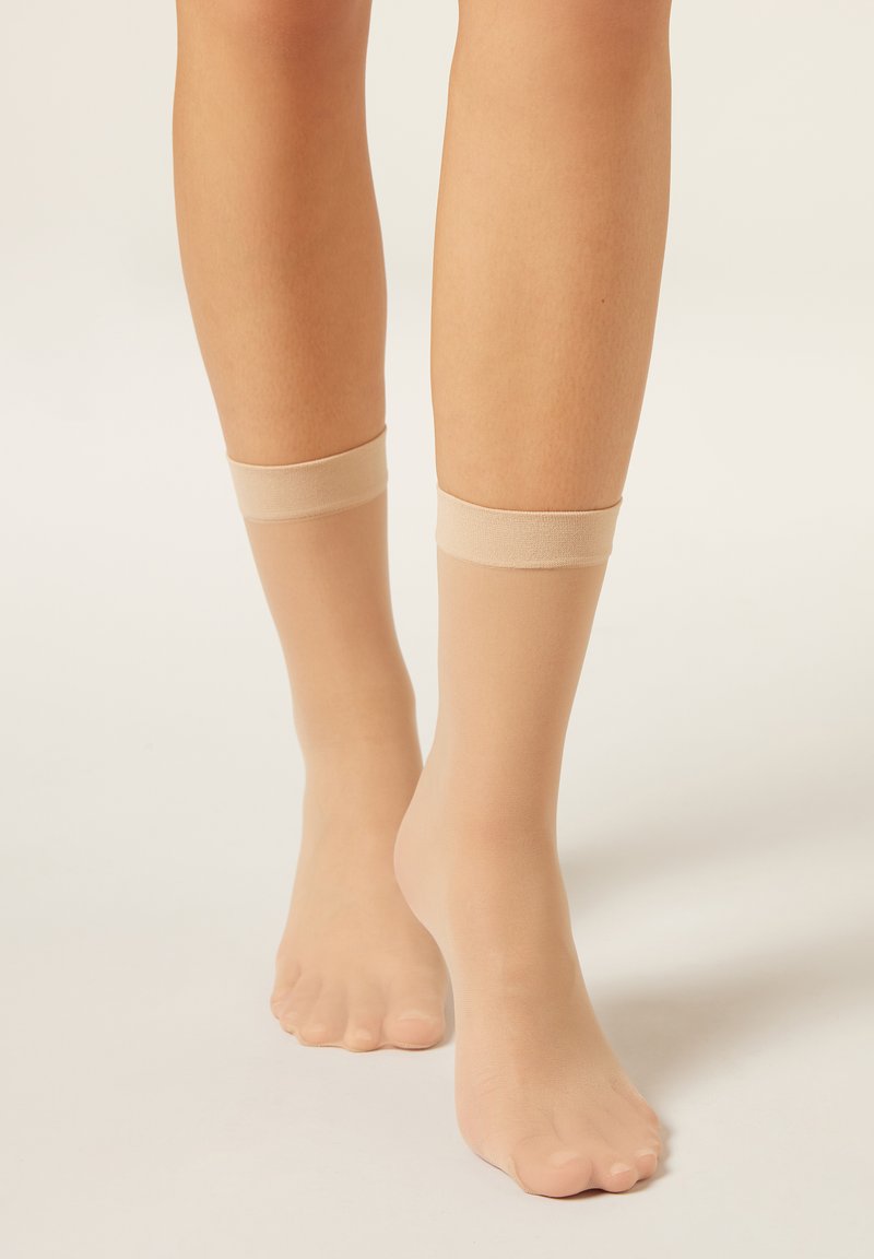 Beige gennemsigtige ankelsokker med en glat tekstur og en solid elastisk kant øverst, der dækker anklerne og en del af fødderne.