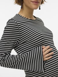 Γυναίκα με μαύρη και λευκή μακρυμάνικη μπλούζα με σχέδιο ζιγκ ζαγκ και μανσέτες με καμπάνα, που κρατά την εγκυμονούσα κοιλιά της, φορώντας σκούρα στρογγυλά σκουλαρίκια.