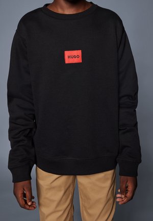 Osoba ubrana w czarną bluzę z długim rękawem z czerwoną naszywką z logo "HUGO" na wysokości klatki piersiowej, w połączeniu z beżowymi spodniami.