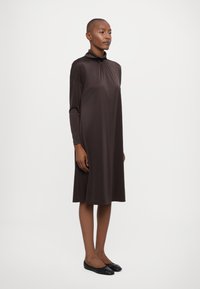 Abito marrone a maniche lunghe con colletto alto, caratterizzato da una forma A-line fluida e una texture morbida, abbinato a delle ballerine nere.