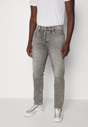 Homme portant un jean gris délavé, un t-shirt blanc et des baskets blanc-gris, se tenant devant un fond clair uni.