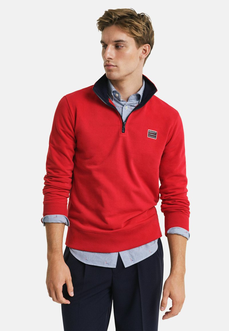 Rode kwart-rits trui met een marineblauwe kraag. Ribgebreide boorden en zoom, gelaagd over een lichtblauw gestreept shirt met kleine patronen. Klassieke pasvorm.