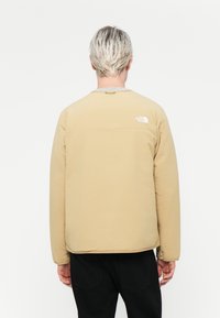 Beige lett jakke med rund hals og lange ermer. Har en liten logo på øvre rygg og glatt tekstur.