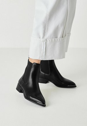 Stivaletti neri in pelle con pannelli laterali elasticizzati indossati con pantaloni bianchi corti su sfondo di pavimento bianco.