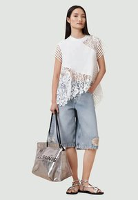 White short-sleeve top with lace and mesh details, paired with distressed denim shorts. Model carries a silver tote bag.