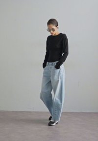 Maglietta nera a maniche lunghe, jeans a gamba larga blu chiaro e scarpe da ginnastica bianche e nere, con occhiali da sole trasparenti, esposti su uno sfondo minimalista.