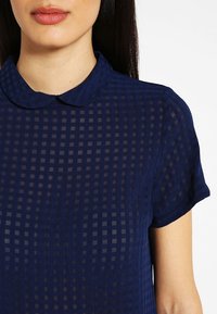 Marinblå kortärmad skjorta med krage. Transparent tyg med ett rutmönster ger ett lätt och texturerat utseende.