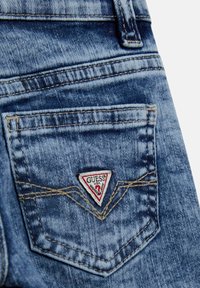 Primo piano di una tasca di jeans blu con un patch triangolare del marchio Guess cucito sopra la cucitura a forma di V.