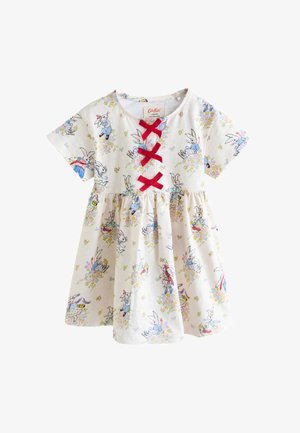 Fată tânără cu păr lung și negru, purtând o rochie crem cu fundițe roșii, șosete albastre și pantofi maro, stând în fața unui tapet floral.