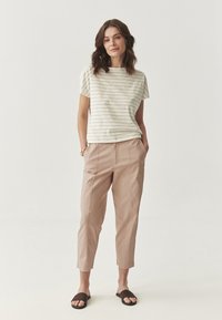 Woman standing with hands in pockets wearing a white striped short-sleeve top, beige cropped pants, and black slip-on sandals.