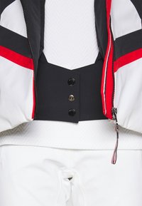 Veste noire raccourcie avec des accents rouges et blancs, comportant quatre boutons noirs et une couche intérieure blanche texturée. Pantalon blanc visible en bas.