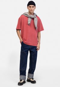 Rdeča oversized majica, mehka tkanina, v kombinaciji z modrimi denim hlačami z vidnimi šivi in zavihanimi manšetami. Oblečena z sivo jopico okoli vratu.