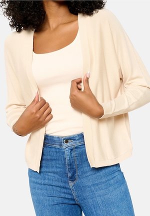 Frau trägt eine beige Strickjacke über einem cremefarbenen Tanktop und blaue hoch taillierte Jeans, hält die Kanten der Strickjacke vor einem schlichten hellen Hintergrund fest.