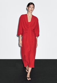 Červené dlouhé šaty s páskem v pase, širokými rukávy a volným střihem. Materiál vypadá lehký a splývavý.
