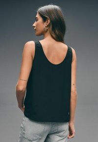 Zwarte mouwloze top met een scoopback ontwerp, gemaakt van een gladde, lichte stof. Gecombineerd met lichtgrijze jeans.