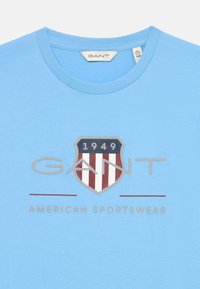 Gaiši zils kokvilnas T-krekls ar vairogu logotipu, sarkaniem un baltajiem svītrainiem, tekstu "GANT" un "1949" pelēkā krāsā virs "AMERICAN SPORTSWEAR."
