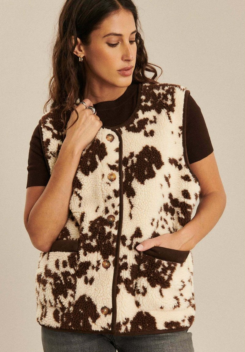 Fleece-Weste mit einem creme- und braunfarbenen Kuhmuster, ausgestattet mit einem Knopfverschluss und zwei seitlichen Taschen. Saubere Nähte und strukturierter Stoff.