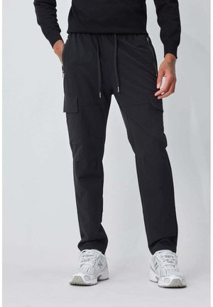 Homme portant un pantalon cargo noir avec poches zippées et à rabat, baskets blanches, debout les mains près des poches sur un fond uni.