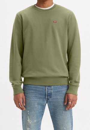 Homme portant un sweat-shirt Levi's vert olive et un jean bleu délavé, debout, les bras relâchés le long du corps, devant un fond uni.