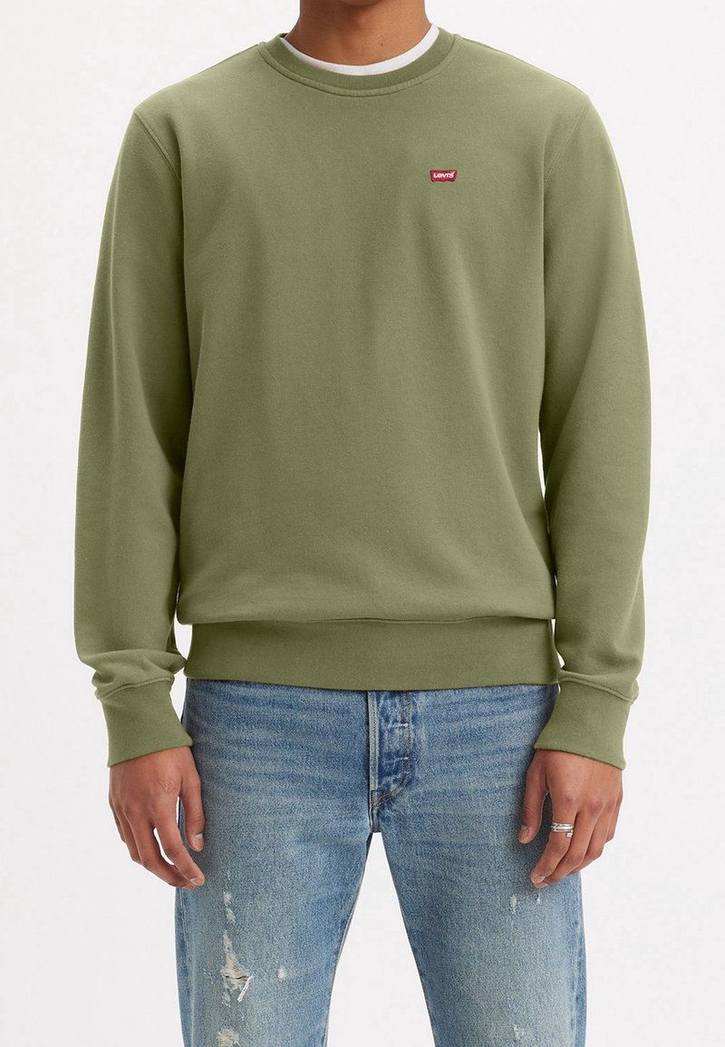 Homme portant un sweat-shirt Levi's vert olive et un jean bleu délavé, debout, les bras relâchés le long du corps, devant un fond uni.