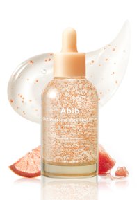 Flacon de sérum avec bouchon compte-gouttes beige contenant un liquide clair avec des perles orange, placé près de tranches de pamplemousse et sur un fond de gel étalé.