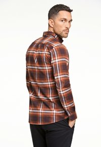 Camicia a quadri marrone con accenti arancioni e bianchi, maniche lunghe e colletto a punta. Il tessuto sembra morbido con una finitura strutturata.