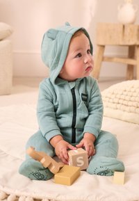 Lyseblå quiltet heldragt med hætte, der har bjørneører og lynlås foran. Babyen sidder på en blød overflade med træklodser.