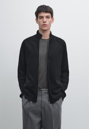 Schwarze Zip-up-Jacke mit einem gerippten Kragen und Schulterdetails, getragen über einem grauen Hemd. Ein Paar graue, maßgeschneiderte Hosen vervollständigt das Outfit.