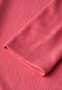 Strickstoff in einem leuchtenden Pink, mit geripptem Muster und umgeschlagenem Rand. Nahtdetails sind durch das Material sichtbar.