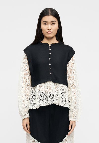 Femme aux longs cheveux noirs portant un haut noir sans manches à boutons sur un chemisier blanc en dentelle avec des motifs floraux, debout devant un fond uni.