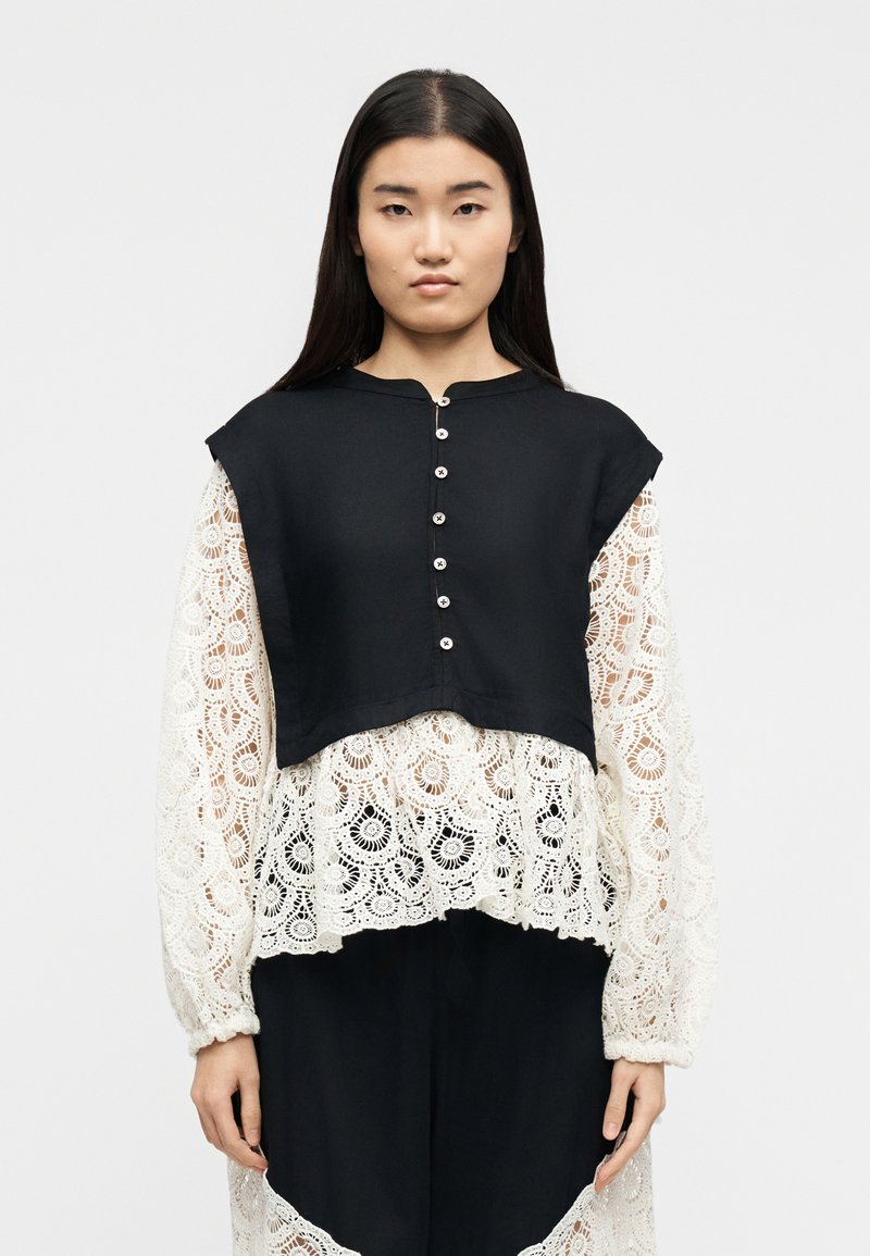 Γυναίκα με μακριά σκουρόχρωμα μαλλιά φοράει μαύ ρο αμάνικο τοπ με κουμπιά πάνω από λευκό δαντελένιο πουκάμισο με λουλουδένια σχέδια, στέκεται μπροστά από απλό φόντο.