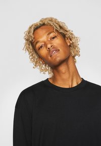 T-shirt noir à manches longues avec un col rond, fabriqué en coton, présentant une coupe décontractée et des détails de couture subtils. Apparence texturée avec une finition douce.