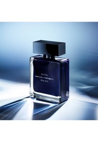 Tapa cuadrada negra en un frasco de perfume de vidrio claro con líquido azul oscuro. La etiqueta dice "para él narciso rodríguez bleu noir." Textura suave.