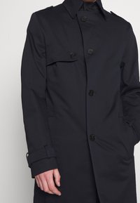 Marinblå trench coat i slätt tyg, med dubbelknäppning på framsidan, svarta knappar och sidofickor. Skräddarsydd passform med justerbara ärmslut.