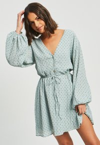 Svetlo modra obleka z V-izrezom, dolgimi napihnjenimi rokavi, sprednjim gumbom in nastavkom na pasu. Ima črne pika in razporek do stegna.