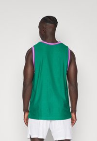 Top sin mangas de malla verde con cuello y agujeros para los brazos en rosa. Presenta un patrón texturizado y un ajuste holgado. Combinado con shorts deportivos blancos.
