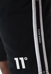 Pantalones cortos negros de algodón, con un logo blanco que dice "11°" en la parte inferior de la pierna y una franja lateral blanca con el texto "ELEVEN DEGREES".
