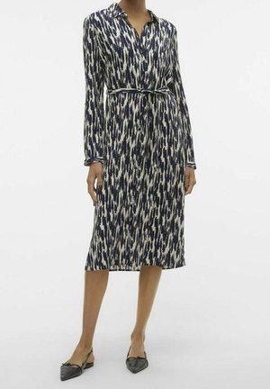 Femme portant une robe boutonnée à manches longues, longueur genou, imprimé abstrait bleu marine et blanc, avec une ceinture nouée à la taille, associée à des ballerines à bride noire et pointe effilée.